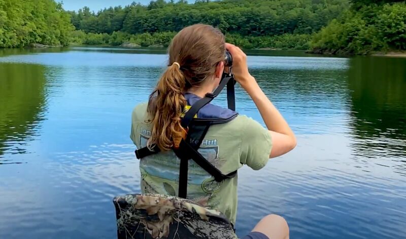 A girl sits in a boat on a lake, looking through binoculars towards the shore, surrounded by calm water and nature